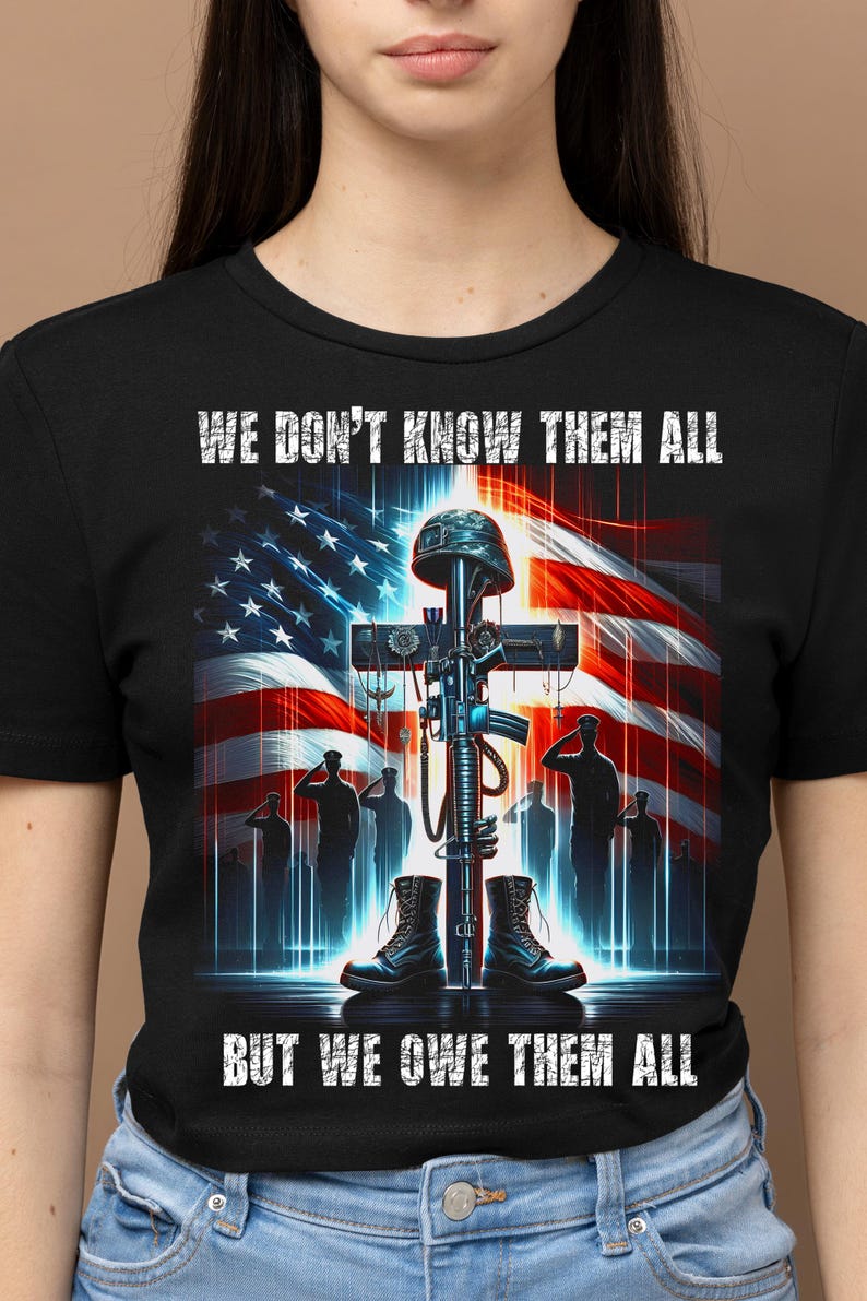 Op de afbeelding: Zwart T-shirt met een afbeelding van een kruis, geweer, helm en laarzen tegen een Amerikaanse vlag. De tekst luidt: "WE DON'T KNOW THEM ALL BUT WE OWE THEM ALL".