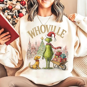 May include: Cream-colored sweatshirt with the word "WHOVILLE" in gold lettering. The graphic features a green character in a Santa hat and a red and white striped scarf, standing next to a dog and a cart of gifts.