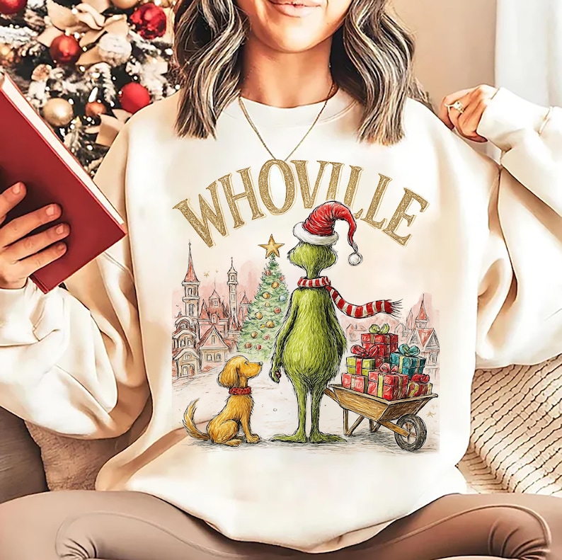May include: Cream-colored sweatshirt with the word "WHOVILLE" in gold lettering. The graphic features a green character in a Santa hat and a red and white striped scarf, standing next to a dog and a cart of gifts.