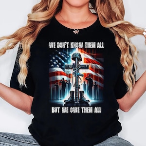 Op de afbeelding: Zwart T-shirt met een afbeelding van een kruis, helm, geweer, laarzen en saluerende soldaten tegen een Amerikaanse vlag. De tekst luidt: "WE DON'T KNOW THEM ALL BUT WE OWE THEM ALL."