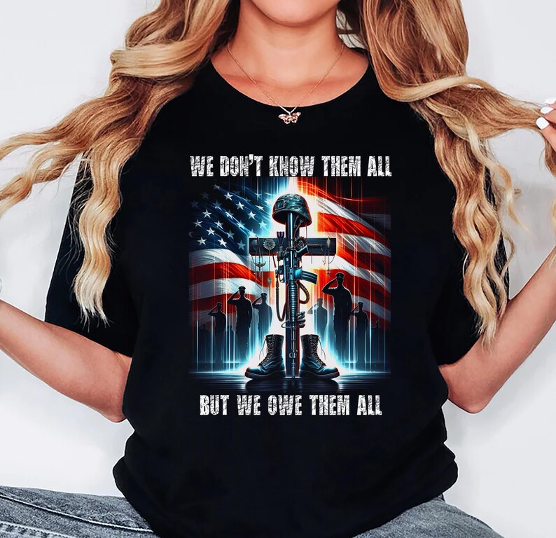 Op de afbeelding: Zwart T-shirt met een afbeelding van een kruis, helm, geweer, laarzen en saluerende soldaten tegen een Amerikaanse vlag. De tekst luidt: "WE DON'T KNOW THEM ALL BUT WE OWE THEM ALL."