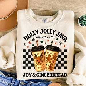 Puede incluir: Sudadera color crema con el texto "HOLLY JOLLY JAVA served with JOY & GINGERBREAD." El diseño presenta dos tazas de café con gráficos festivos, incluyendo un reno y hombres de jengibre, sobre un fondo a cuadros blanco y negro.