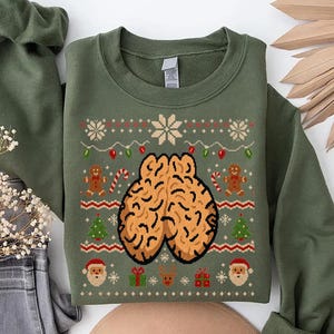 Puede incluir: Una sudadera de cuello redondo en color verde oliva con un patrón festivo. El diseño incluye un gráfico central de un cerebro, rodeado de figuras de pan de jengibre, bastones de caramelo, árboles de Navidad, caras de Santa Claus, renos y regalos.