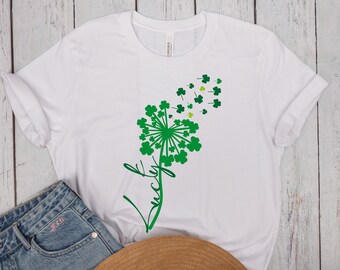 Puede incluir: Camiseta blanca con un diseño de trébol verde que se asemeja a un diente de león. El diseño dice "Lucky" en cursiva.