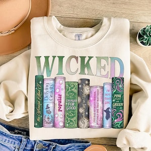 Puede incluir: Sudadera color crema con la palabra "WICKED" en letras verdes e iridiscentes. Debajo, una fila de gráficos coloridos con temática de libros con textos como "Defying Gravity" y "Popular". Un sombrero y unos vaqueros están en el fondo.