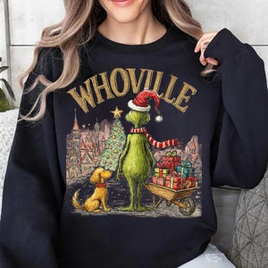 May include: Black sweatshirt with a graphic featuring the word "WHOVILLE" in gold lettering. The design includes a green character wearing a Santa hat and scarf, a dog, a Christmas tree, and a cart of gifts. The background depicts a town scene.