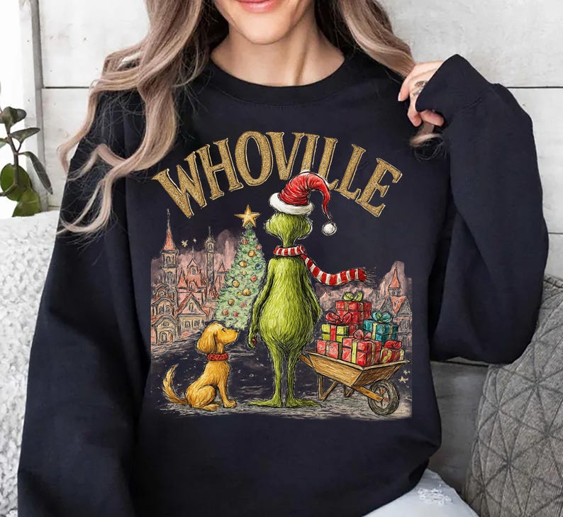 May include: Black sweatshirt with a graphic featuring the word "WHOVILLE" in gold lettering. The design includes a green character wearing a Santa hat and scarf, a dog, a Christmas tree, and a cart of gifts. The background depicts a town scene.