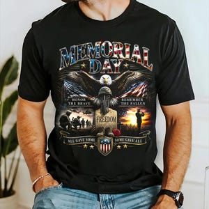 May include: Black t-shirt with a Memorial Day graphic. The design features an eagle, American flags, and the text "Memorial Day." Additional text includes "Honor the Brave," "Remember the Fallen," and "Freedom."