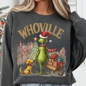 May include: Dark gray sweatshirt featuring a whimsical illustration of a green character in a Santa hat and scarf, standing next to a dog and a Christmas tree. The word "WHOVILLE" is arched across the top. A wheelbarrow of gifts is also present.