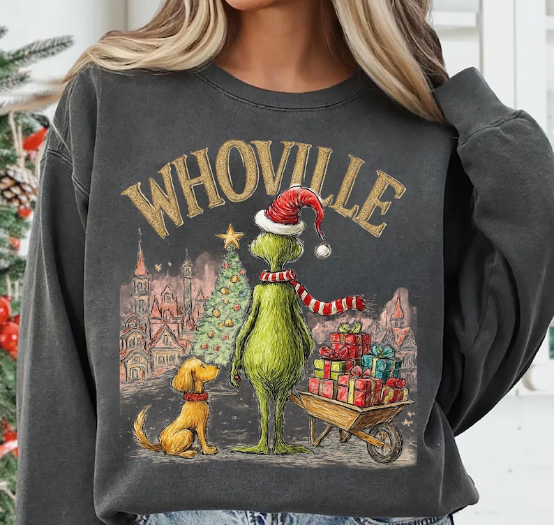 May include: Dark gray sweatshirt featuring a whimsical illustration of a green character in a Santa hat and scarf, standing next to a dog and a Christmas tree. The word "WHOVILLE" is arched across the top. A wheelbarrow of gifts is also present.