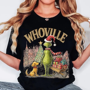May include: Black t-shirt featuring a Christmas-themed illustration. The design includes the text "WHOVILLE" in gold lettering, a green character wearing a Santa hat and scarf, a Christmas tree, a dog, and a wheelbarrow filled with gifts.
