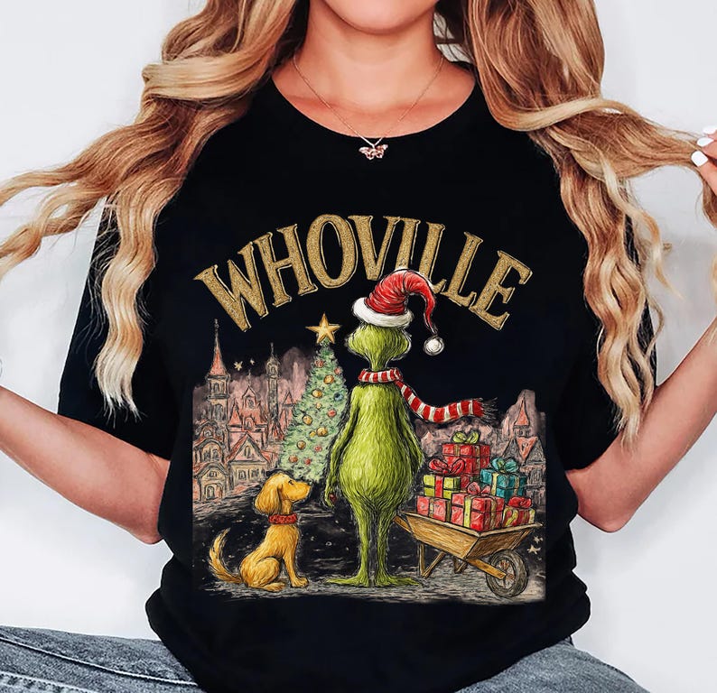 May include: Black t-shirt featuring a Christmas-themed illustration. The design includes the text "WHOVILLE" in gold lettering, a green character wearing a Santa hat and scarf, a Christmas tree, a dog, and a wheelbarrow filled with gifts.