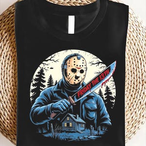May include: Black t-shirt featuring a graphic of a masked figure holding a bloody knife with the text "FRIDAY THE 13TH". The design includes a cabin, trees, and a full moon, creating a horror movie theme.