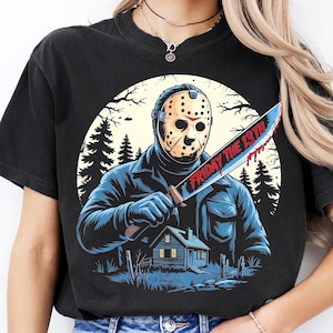 May include: Black t-shirt featuring a graphic of a masked figure holding a bloody knife with the text "FRIDAY THE 13TH". The design includes a house, trees, and a full moon, creating a horror movie theme.