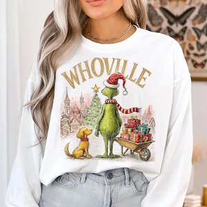 May include: White long-sleeve sweatshirt featuring a whimsical illustration of the Grinch in a Santa hat and scarf, with a dog and gifts. The word "WHoville" is printed above the Grinch. A Christmas tree and buildings are in the background.