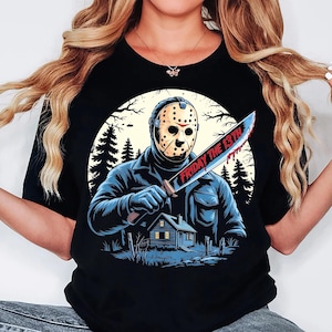 May include: Black t-shirt featuring a graphic of a masked figure holding a bloody knife with the text "FRIDAY THE 13TH". The design includes a house, trees, and a full moon in the background. The shirt is a crew neck.