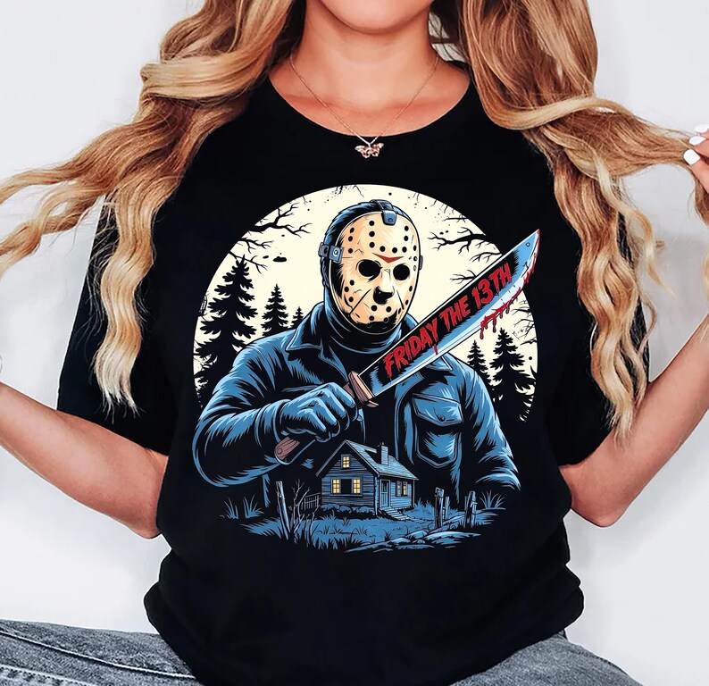 May include: Black t-shirt featuring a graphic of a masked figure holding a bloody knife with the text "FRIDAY THE 13TH". The design includes a house, trees, and a full moon in the background. The shirt is a crew neck.