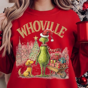 May include: Red sweatshirt featuring a graphic of the Grinch in a Santa hat and scarf, with a dog and a cart of gifts. The word "Whoville" is printed above the Grinch in gold lettering. A Christmas tree and buildings are in the background.