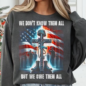 Op de afbeelding: Donkergrijze sweatshirt met een Amerikaans vlagontwerp, een kruis, helm, laarzen en een geweer. De tekst luidt "WE DON'T KNOW THEM ALL BUT WE OWE THEM ALL". Het ontwerp is een eerbetoon aan militair personeel.