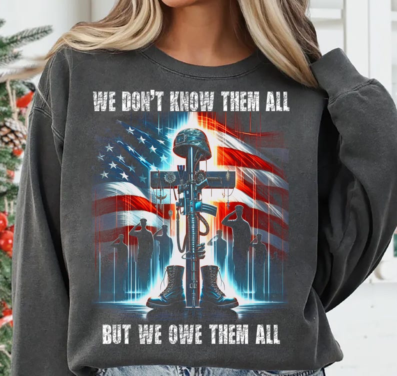 Op de afbeelding: Donkergrijze sweatshirt met een Amerikaans vlagontwerp, een kruis, helm, laarzen en een geweer. De tekst luidt "WE DON'T KNOW THEM ALL BUT WE OWE THEM ALL". Het ontwerp is een eerbetoon aan militair personeel.