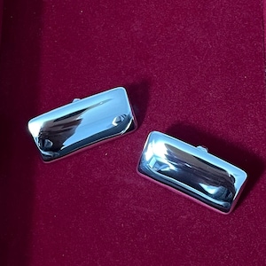 May include: Two polished, rectangular silver cufflinks with rounded edges. The cufflinks reflect light, creating a shiny appearance. They are displayed on a deep red velvet surface.