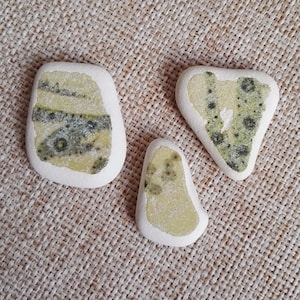 May include: Three smooth, white stones with a green and yellow pattern. The stones are arranged in a triangular shape on a beige fabric background.