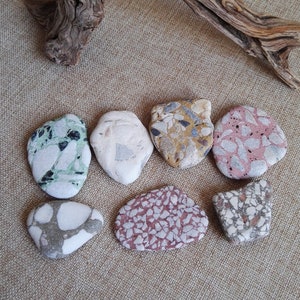 May include: Eight smooth, round, colorful stones with a speckled pattern. The stones are arranged in a semi-circle on a beige fabric background.