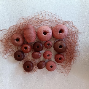 Op de afbeelding: Een verzameling van verschillende maten, roodbruine houten kralen en boeien, gerangschikt op een stuk bruin net. De kralen variëren in vorm, van rond tot langwerpig, sommige met gaten. De achtergrond is een getextureerd wit oppervlak.