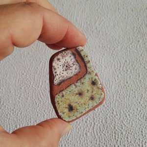 May include: A small, irregularly shaped ceramic tile with a brown, green, and white glaze. The glaze has a speckled pattern with brown and white dots.