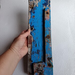 Op de afbeelding: Een verweerd houten plank met blauwe verf, met tekenen van slijtage. De plank heeft een metalen scharnier eraan bevestigd, en er zijn verschillende schroeven zichtbaar. De plank is een gered stuk hout, perfect voor knutselen of decoreren.