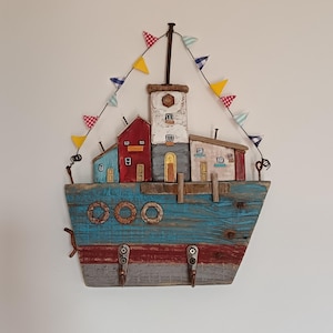 Può includere: Un'applique da parete in legno a forma di barca, con case in miniatura e un faro. La barca è dipinta di blu e rosso, con tre ganci in metallo. Striscioni colorati sono appesi sopra le case. Le case sono dipinte di rosso, blu, bianco e beige.