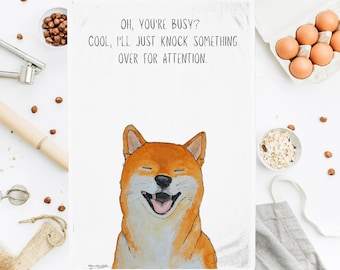 Puede incluir: Una toalla de cocina blanca con un dibujo animado de un perro Shiba Inu con una expresión feliz. El texto en la toalla dice "OH, YOU'RE BUSY? COOL, I'LL JUST KNOCK SOMETHING OVER FOR ATTENTION."