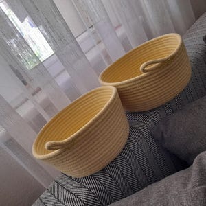 May include: Two light yellow rope baskets with small handles. The baskets sit on a grey and white patterned surface, with grey pillows and a sheer curtain in the background. The baskets are made of tightly woven rope.
