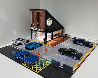 Może przedstawiać: Miniaturowy model sklepu Starbucks z drive-thru i parkingiem. Sklep jest wykonany z drewna i ma zielono-czarne logo. Na parkingu stoi kilka miniaturowych samochodów, w tym niebieski Lamborghini, biały samochód z naklejką "LOWK" i czarny samochód z naklejką "LOWK".