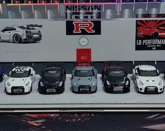 May include: A display case with a collection of seven miniature Nissan Skyline GT-R cars. The cars are painted in various colors, including blue, white, black, and gray. The case has a white background with the words "Japanese Muscle" and "LB Performance" printed on it.