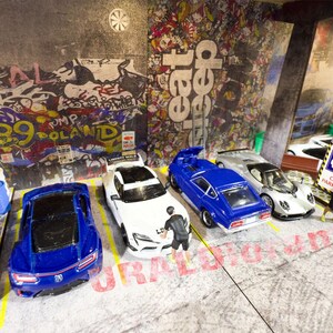 May include: A collection of toy cars parked in a garage setting. The cars are a white and blue sports car, a blue and white sports car, a silver sports car, a white sports car, and a blue sports car. The garage has a concrete floor and a graffiti-covered wall.