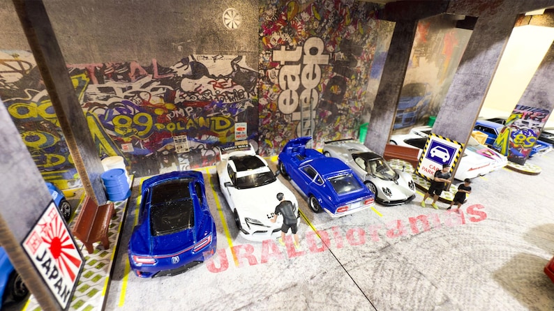 May include: A collection of toy cars parked in a garage setting. The cars are a white and blue sports car, a blue and white sports car, a silver sports car, a white sports car, and a blue sports car. The garage has a concrete floor and a graffiti-covered wall.