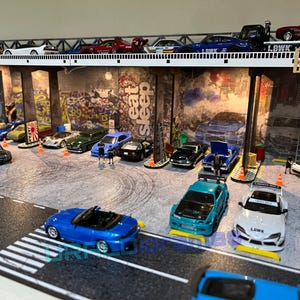 May include: A model car diorama set with a bridge and a variety of cars, including a blue convertible, a white car with a black stripe, and a red car with a black stripe. The diorama is set against a backdrop of a graffiti-covered wall with the words "Eat Sleep" written on it.