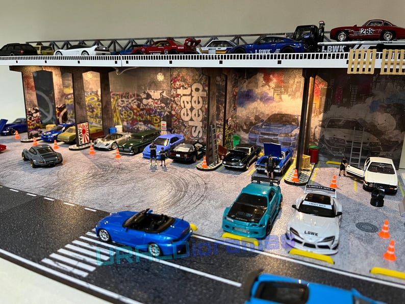 May include: A model car diorama set with a bridge and a variety of cars, including a blue convertible, a white car with a black stripe, and a red car with a black stripe. The diorama is set against a backdrop of a graffiti-covered wall with the words "Eat Sleep" written on it.