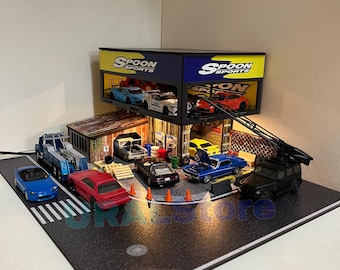 May include: A miniature model of a garage with a sign that reads "Spoon Sports" and several miniature cars inside. The garage is lit with LED lights and has a black base with white lines.