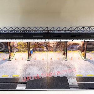 May include: A model of a city street scene with a graffiti-covered building and a black metal overpass. The scene is lit with white lights and includes a black asphalt road with white lane markings.