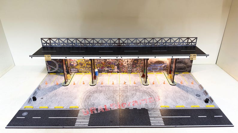 May include: A model of a city street scene with a graffiti-covered building and a black metal overpass. The scene is lit with white lights and includes a black asphalt road with white lane markings.