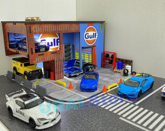 May include: A miniature model of a garage with a Gulf gas station sign, a yellow truck, and several sports cars. The garage has a red brick exterior and a blue and white interior. The cars are all in different colors and styles. The model is made of wood and plastic.
