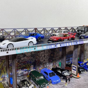 May include: A collection of miniature toy cars, including a white Honda NSX, a red and white Nissan Skyline, a blue Honda Civic, and a black Nissan 240SX, are displayed on a model bridge with a graffiti backdrop.
