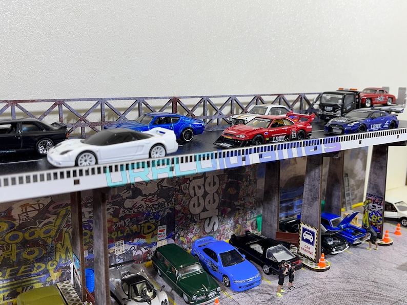 May include: A collection of miniature toy cars, including a white Honda NSX, a red and white Nissan Skyline, a blue Honda Civic, and a black Nissan 240SX, are displayed on a model bridge with a graffiti backdrop.