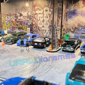 May include: A collection of miniature toy cars in a diorama setting. The cars are a mix of colors, including blue, green, black, and white. The diorama features a brick wall with graffiti and a street scene. The cars are arranged in a line, with some of them having their hoods open. The diorama is a popular hobby for collectors and enthusiasts.