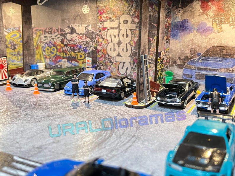 May include: A collection of miniature toy cars in a diorama setting. The cars are a mix of colors, including blue, green, black, and white. The diorama features a brick wall with graffiti and a street scene. The cars are arranged in a line, with some of them having their hoods open. The diorama is a popular hobby for collectors and enthusiasts.