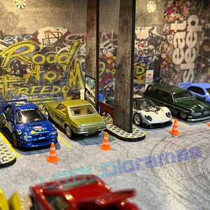 May include: A collection of toy cars parked in a garage setting. The cars are a blue Subaru Impreza, a green station wagon, a white Pagani Zonda, a light blue Honda Civic, and a yellow car. The garage has a concrete floor and a graffiti wall with the words "Road to Freedom".