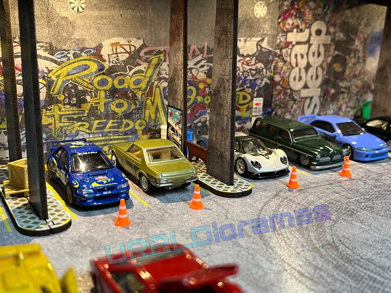 May include: A collection of toy cars parked in a garage setting. The cars are a blue Subaru Impreza, a green station wagon, a white Pagani Zonda, a light blue Honda Civic, and a yellow car. The garage has a concrete floor and a graffiti wall with the words "Road to Freedom".