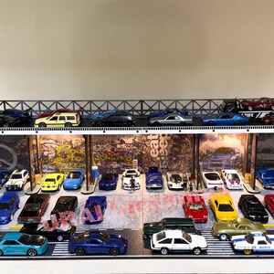 May include: A multi-level parking garage display with a variety of miniature cars in different colors, including red, blue, yellow, black, and white. The cars are arranged in rows on the display, with some cars on the upper level and others on the lower level. The display is made of wood and metal and has a graffiti-style backdrop.